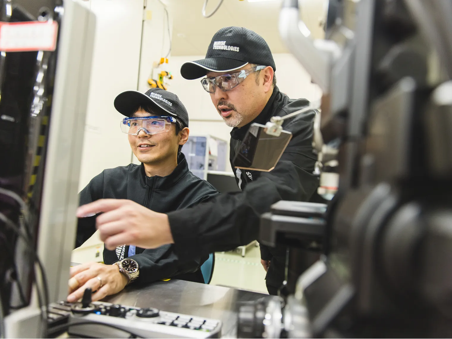 研究施設で設備を操作しながら議論する長里と徳田