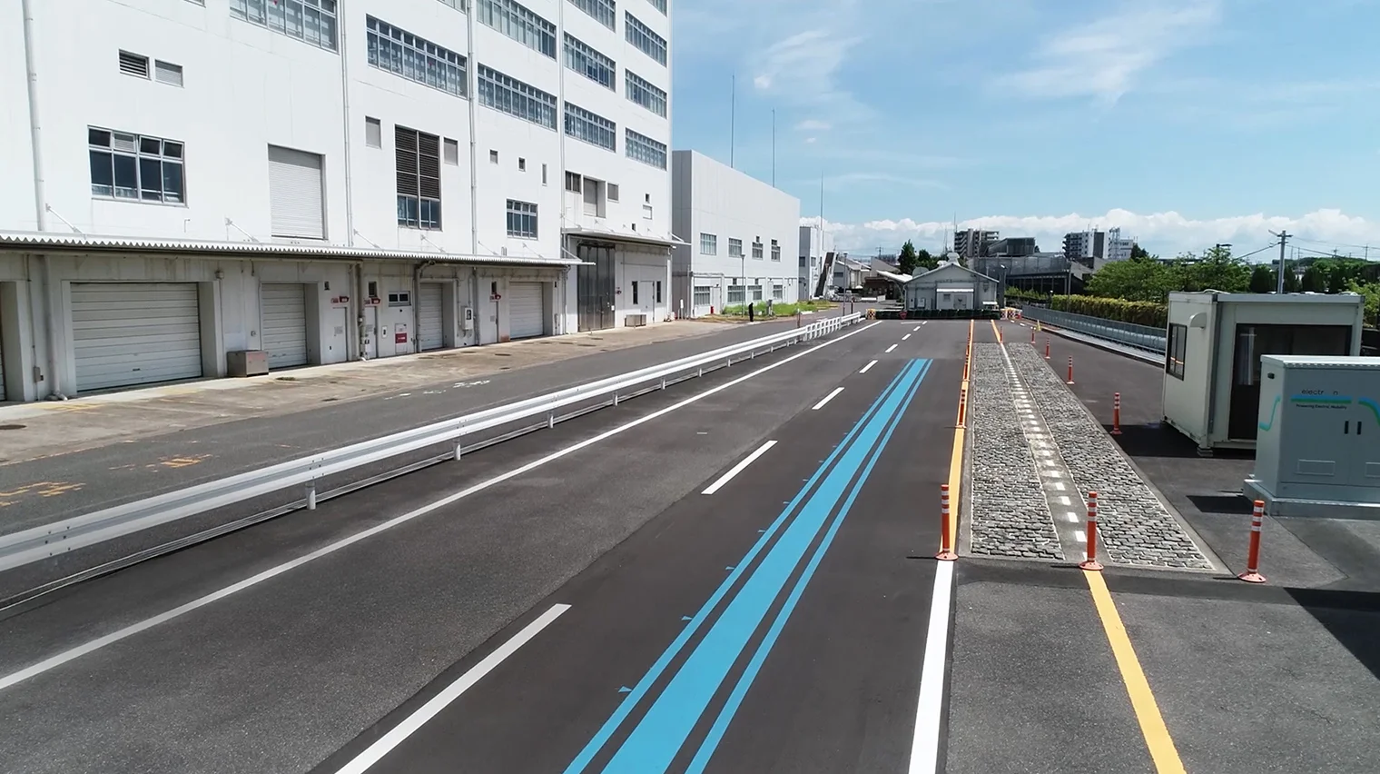 An outdoor test course for demonstration trials of wireless power transfer while driving. A power-supply lane marked by a blue line on the asphalt is visible, along with a factory, a control building, and power-supply equipment.