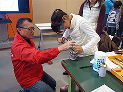 写真は、真ちゅう板の不要な部分を弓のこで切る作業。アドバイスをもらいながら、直角に刃を当て切断している様子