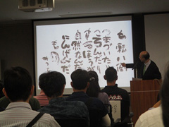 写真は、相田かずひと氏が、相田みつをの書「しちてんばっとう」を指さして、講演している様子