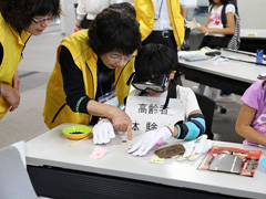 写真は、来場者の子供が、ゴーグルをつけて高齢者疑似体験をする様子