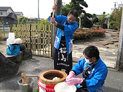 写真は、青い法被を着た男性が杵を高く振り上げて餅をついている様子