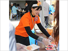 写真は、オレンジいろのTシャツを着た若い女性が来場者とやり取りをしながら餅を渡している様子